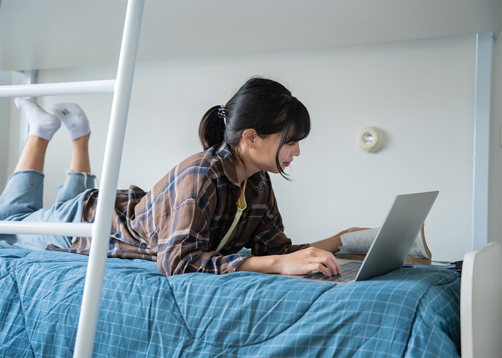student studying with book and laptop on bed in dorm room