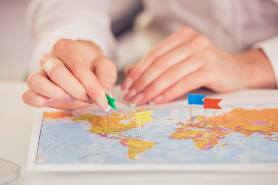World map sitting on table with person sticking flag pins in it in various locations