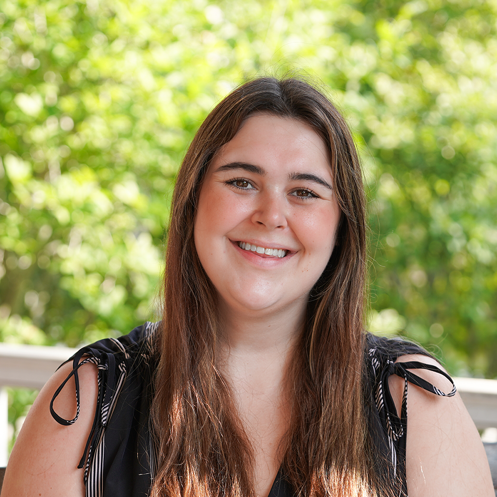 Young woman with long brown hair, smiling outdoors.