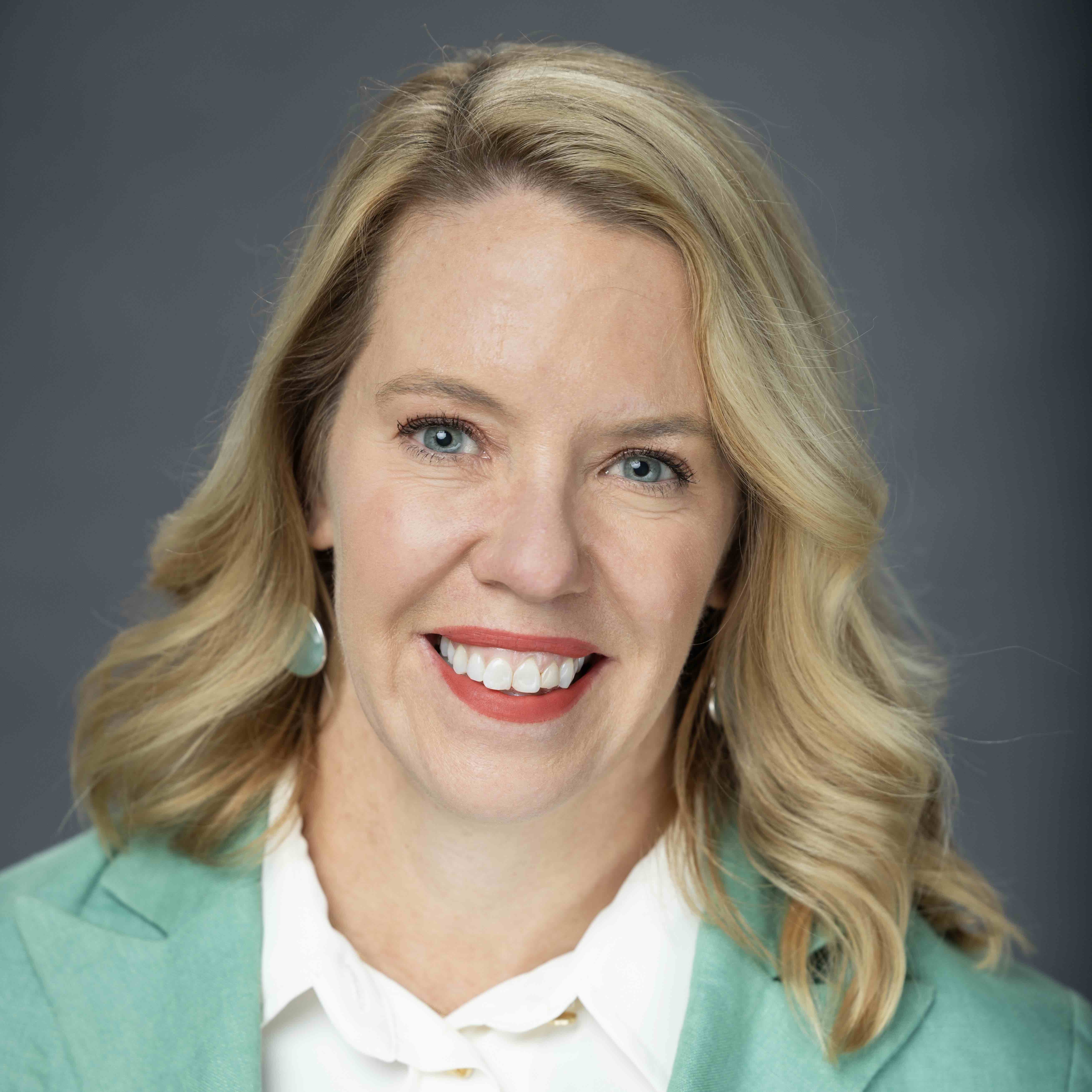 Photograph of a smiling blonde woman with blue eyes wearing a teal suit jacket.