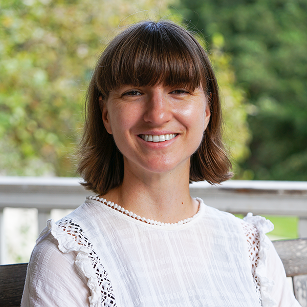 Young woman with short brown hair and bangs, smiling, wearing a white lace-trimmed blouse outdoors.