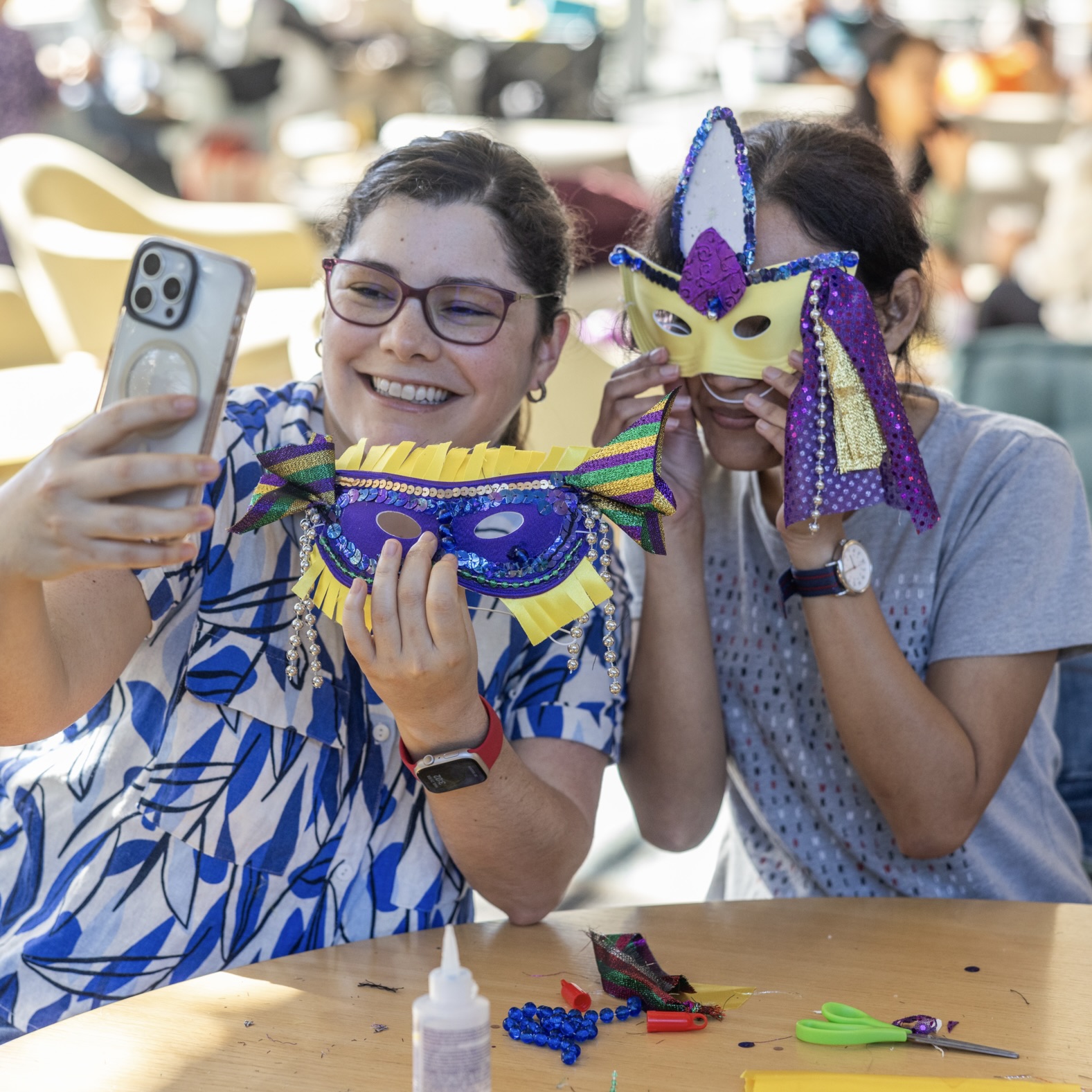 Two women make Mardi Gras masks, one taking a selfie.