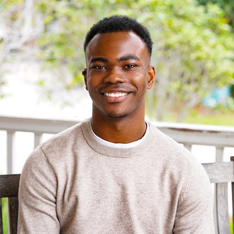 Young Black man smiling in a beige sweater with an outdoor background.
